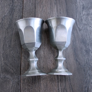 May include: Two silver goblets with a fluted design and a rounded base. The goblets are made of pewter and have a shiny finish.