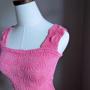 May include: A bright pink dress with a square neckline and textured floral pattern. The dress is displayed on a white mannequin, showcasing the intricate details of the fabric and design. The dress has a fitted bodice and shoulder straps.