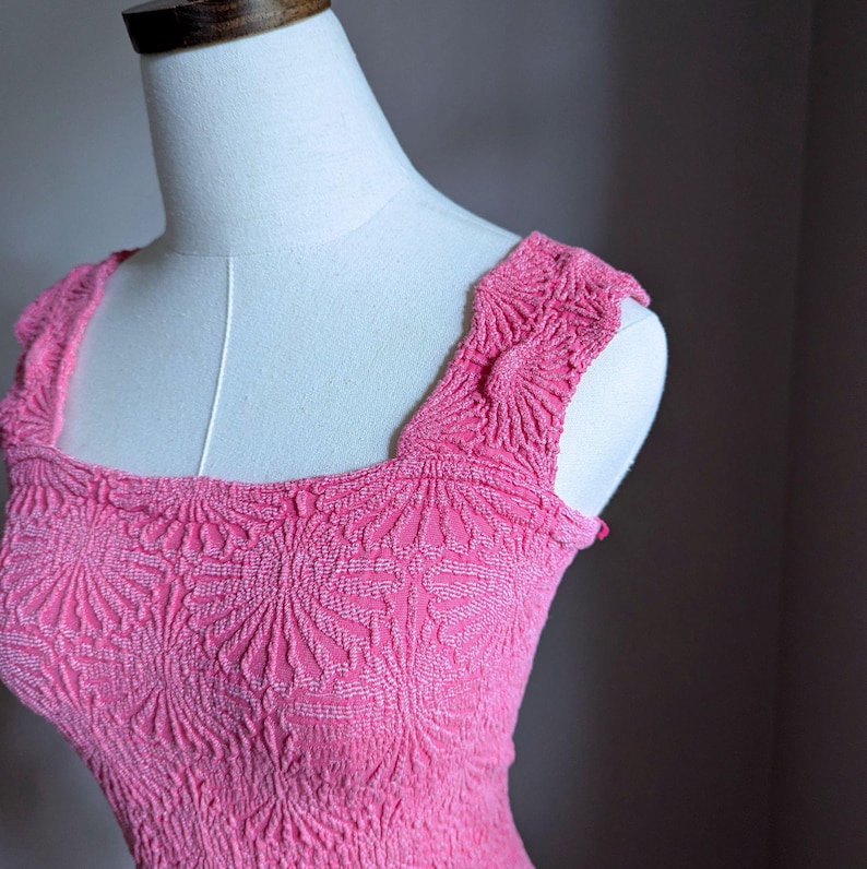 May include: A bright pink dress with a square neckline and textured floral pattern. The dress is displayed on a white mannequin, showcasing the intricate details of the fabric and design. The dress has a fitted bodice and shoulder straps.