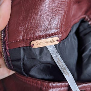 May include: Close-up of a burgundy leather jacket with a Free People brand tag. The tag is made of metal and has a brown finish.