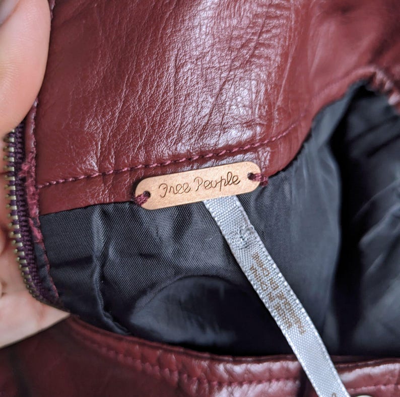 May include: Close-up of a burgundy leather jacket with a Free People brand tag. The tag is made of metal and has a brown finish.