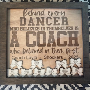 May include: Wooden framed sign with the text "Behind every DANCER who believes in themselves is A COACH who believed in them first." Includes names on bow-shaped cutouts. The sign is for Coach Layla and the Shockers 2025.