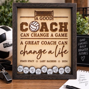 May include: A rectangular wooden shadow box frame with a light wood backing featuring laser-engraved text that reads A GOOD COACH CAN CHANGE A GAME A GREAT COACH CAN CHANGE A LIFE, COACH STACY, LADY RAIDERS, 2024, and nine small volleyball tokens.