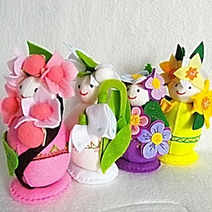 Pode incluir: Quatro bonecas de flores de feltro em rosa, branco, roxo e amarelo. Cada boneca tem um rosto sorridente e é adornada com flores de feltro e folhas verdes. As bonecas estão em uma superfície branca.