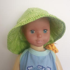 Può includere: Una bambola con un cappello da sole a pois verdi e bianchi con una canotta blu con fiori ricamati. La bambola ha la pelle marrone e gli occhi azzurri.