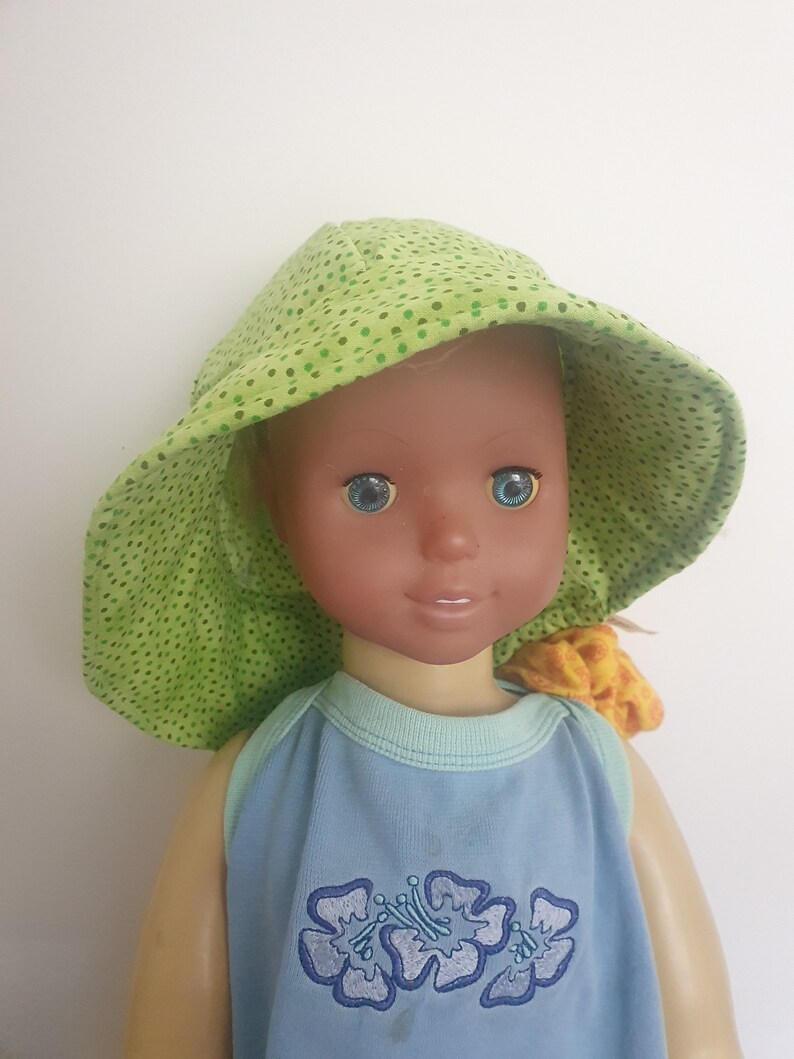 K&ouml;nnte beinhalten: Eine Puppe mit einem gr&uuml;n-wei&szlig;en gepunkteten Sonnenhut und einem blauen Tanktop mit gestickten Blumen. Die Puppe hat braune Haut und blaue Augen.