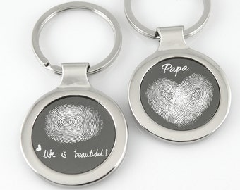 Puede incluir: Dos llaveros plateados con etiquetas redondas de metal grabadas. Una etiqueta presenta un diseño de huella digital en forma de corazón con la palabra "Papa" encima. La otra etiqueta tiene un diseño de huella digital y el texto "life is beautiful".