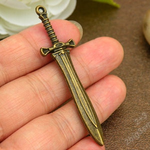 May include: A bronze-colored sword charm with intricate details. The sword has a pointed tip and a cross guard. The charm is perfect for adding a touch of medieval flair to jewelry or crafts.
