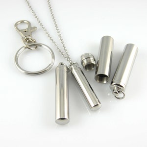 May include: A silver necklace with a small silver cylinder pendant. The pendant can be opened and closed, and there is a matching silver keychain with a cylinder charm.