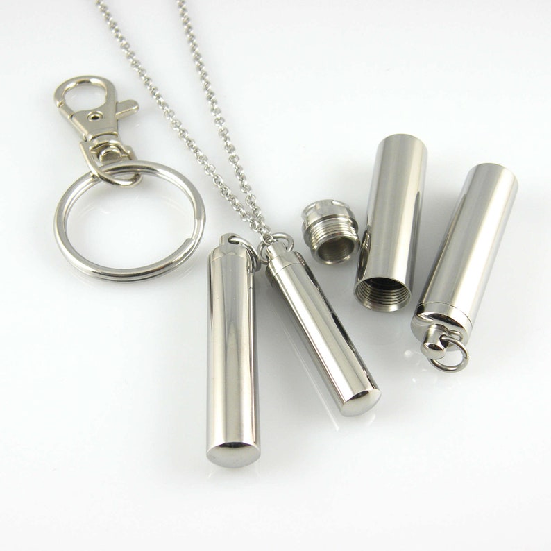 May include: A silver necklace with a small silver cylinder pendant. The pendant can be opened and closed, and there is a matching silver keychain with a cylinder charm.