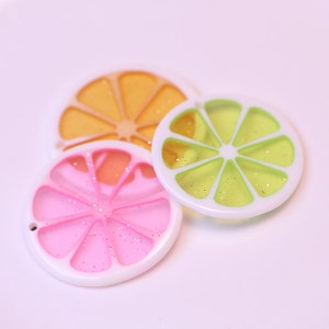 May include: Three colorful plastic citrus fruit slices with glitter inside. The slices are orange, pink, and green.