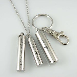 May include: Two silver cylindrical pendants with engraved text. One pendant is on a silver chain necklace, the other is attached to a silver key ring. The necklace pendant reads "Angelia Kevin" with a heart symbol. The key ring pendant reads "love you" with a heart symbol. The third pendant reads "Add your text or logo here".