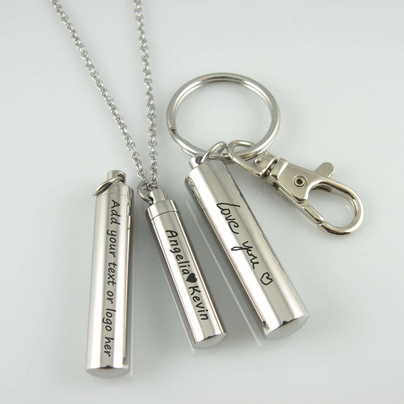 May include: Two silver cylindrical pendants with engraved text. One pendant is on a silver chain necklace, the other is attached to a silver key ring. The necklace pendant reads "Angelia Kevin" with a heart symbol. The key ring pendant reads "love you" with a heart symbol. The third pendant reads "Add your text or logo here".