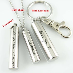 May include: Three silver metal cylindrical lockets with different engravings. The locket on the left says "Add your text or logo here". The middle locket says "Angelia Kevin" with a small heart symbol. The locket on the right says "Love you" with a heart symbol. The lockets are shown with a chain and a keychain.