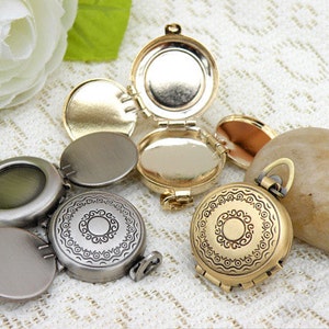 Op de afbeelding: Twee antieke medaillon hangers, een zilveren en een gouden. Beide hebben een cirkelvormig ontwerp met een decoratieve rand en een scharnierend deksel. Het zilveren medaillon heeft een glad interieur, terwijl het gouden medaillon een bloemmotief heeft.