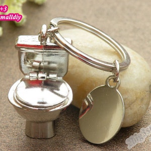 Pode incluir: Um porta-chaves de metal prateado com um pendente de sanita em miniatura e uma pequena etiqueta de metal oval e lisa presa a um anel circular. O pendente de sanita inclui uma tampa e um assento articulados, com o texto 2+3 2big3smalldiy.
