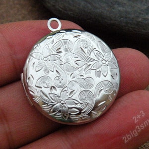 May include: A silver round locket with a floral and swirl pattern. It has a small loop for a chain. The locket is open and empty. The locket is held in a person's hand. The locket is a piece of jewellery.