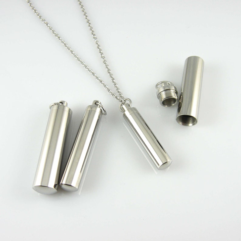 May include: Three silver cylindrical pendants with a silver chain. Two pendants are closed and one is open, revealing an empty interior.