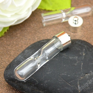 May include: A clear glass hourglass pendant with a silver cap. The hourglass is filled with a small amount of sand and is resting on a black stone.