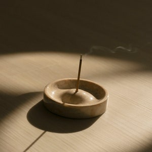 May include: A beige marble incense holder with a single incense stick burning and releasing smoke. The holder is on a light wood surface.