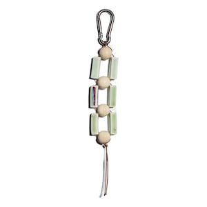 Peut inclure: Un porte-clés décoratif avec un mousqueton argenté. Le porte-clés présente une série de carreaux de céramique rectangulaires vert clair et des perles sphériques de couleur crème, reliés par un cordon marron. Une petite gland pend en bas.