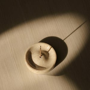 May include: A white ceramic incense holder with a small hole in the center for an incense stick. The holder is round and has a slightly raised edge.