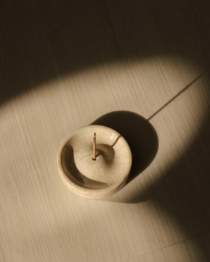 May include: A white ceramic incense holder with a small hole in the center for an incense stick. The holder is round and has a slightly raised edge.
