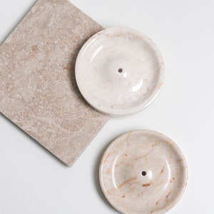May include: Two round, white marble dishes with a small hole in the center. The dishes are on a beige marble surface.