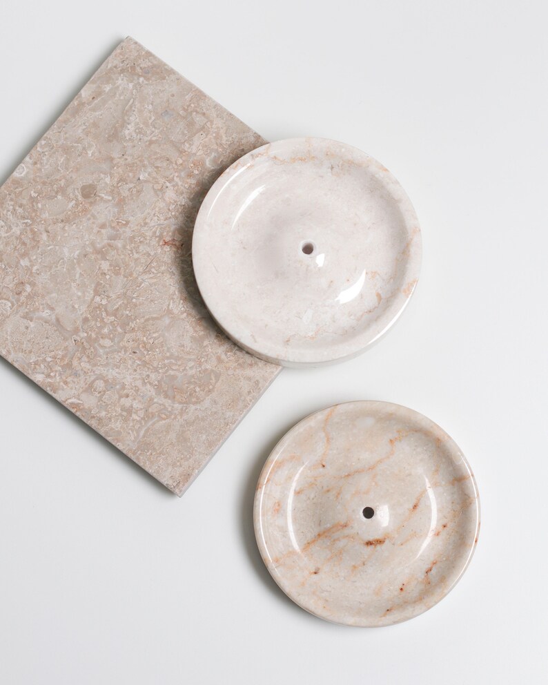 May include: Two round, white marble dishes with a small hole in the center. The dishes are on a beige marble surface.