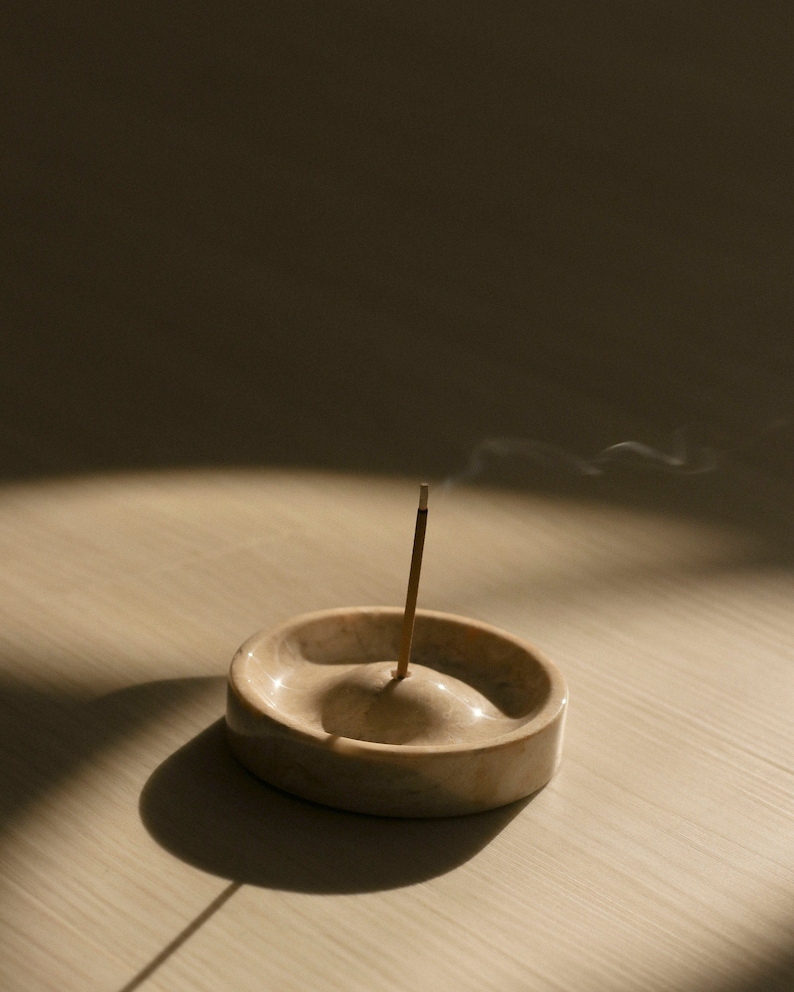 May include: A beige marble incense holder with a single incense stick burning and releasing smoke. The holder is on a light wood surface.