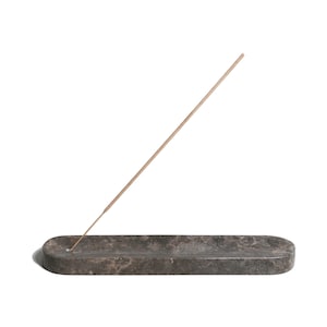 Könnte beinhalten: Ein grauer Marmor-Räucherstäbchenhalter mit einem brennenden Räucherstäbchen. Der Halter ist rechteckig und hat eine leicht abgerundete Kante.