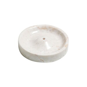 May include: White marble incense holder with a small hole in the center. The holder has a slightly raised edge and a smooth, polished surface.