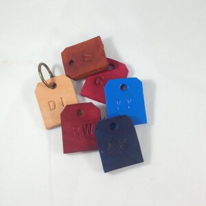 Op de afbeelding: Zes leren sleutelhangers met verschillende kleuren leer en initialen. De sleutelhangers zijn in de volgende kleuren: bruin, beige, rood, blauw en een lichtere tint rood. De initialen op de sleutelhangers zijn: DI, RW, AK, KK, M en G.