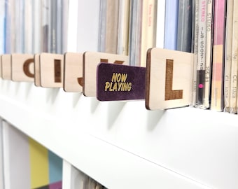 Puede incluir: Separadores de discos de vinilo de madera con letras grabadas y un separador morado "NOW PLAYING". Los separadores se colocan entre los discos de vinilo en un estante blanco, creando una colección de música organizada.