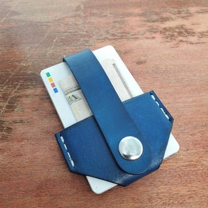 Könnte beinhalten: Ein blauer Lederkartenhalter mit weißen Nähten und einem silbernen Knopf. Der Halter hält eine Kreditkarte und einen Dollar-Schein.