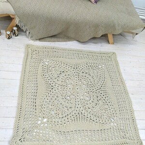 May include: A beige crocheted square rug with a detailed, intricate pattern in the center. The rug is laid out on a white wooden floor.