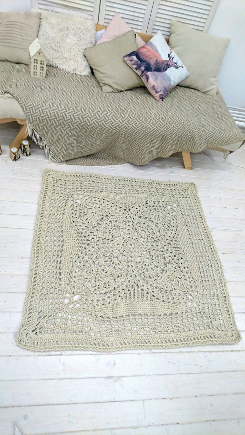 May include: A beige crocheted square rug with a detailed, intricate pattern in the center. The rug is laid out on a white wooden floor.