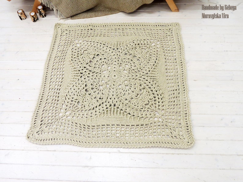 May include: A square, crocheted rug in a light beige color with a detailed, intricate pattern. The rug has a border around the edge and a larger, more detailed pattern in the center.
