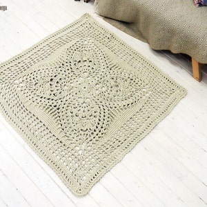 May include: A beige crocheted rug with a square shape and a detailed, intricate pattern. The rug is laid out on a white wooden floor.