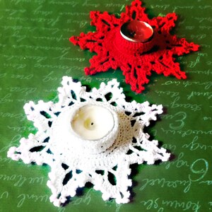 May include: Two crocheted snowflake candle holders, one white and one red, with a tealight candle in each. The candle holders are on a green surface.