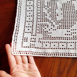 May include: Close-up of a white, crocheted doily with a detailed openwork pattern. The doily has a scalloped edge and is partially held by a hand. The intricate design suggests a handmade, vintage aesthetic, suitable for home decor.