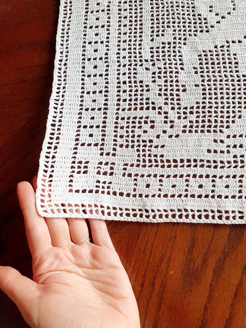 May include: Close-up of a white, crocheted doily with a detailed openwork pattern. The doily has a scalloped edge and is partially held by a hand. The intricate design suggests a handmade, vintage aesthetic, suitable for home decor.