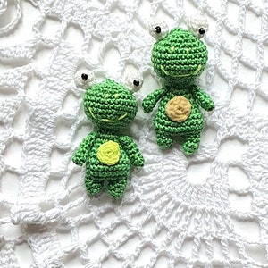 May include: Two small green crocheted frog plushies with white eyes and yellow spots on a white crocheted doily.