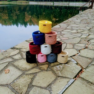 May include: A stack of ten colorful crocheted baskets in various colors, including yellow, blue, red, pink, white, and black. The baskets are arranged in a pyramid shape on a stone surface.