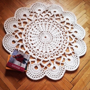 May include: A cream-coloured, handmade crochet doily with a scalloped edge. The circular design features intricate openwork patterns. A stack of books rests on the wooden floor beneath the doily.