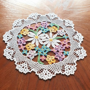May include: A handmade, white crochet doily with a floral design. The center features large white daisies with yellow centers, surrounded by smaller flowers in shades of pink, yellow, blue, and purple. The doily has a scalloped edge.