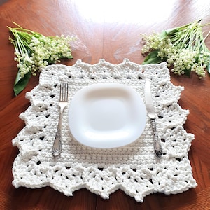 Op de afbeelding: Een wit gehaakt placemat met een geschulpte rand, een wit bord en bestek zijn op een houten tafel geplaatst. Takjes witte bloemen met groene bladeren zijn aan weerszijden van de placemat geplaatst.