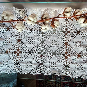 Puede incluir: Doily de crochet blanco con un delicado patrón floral, con una rama de bolas de algodón en la parte superior.