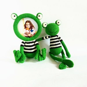 May include: Two green crocheted frog photo frames. The larger frog has a round frame with a picture of a child inside. Both frogs have black and white striped shirts.