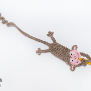May include: A brown crocheted monkey bookmark with pink facial features and a yellow banana in its hand. The monkey's tail is curled into a spiral.